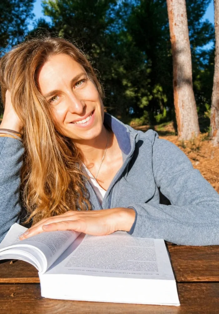 Mujer sonriente con cabello ondulado, sentada al aire libre, leyendo un libro en una mesa de madera rodeada de árboles. La luz del sol ilumina su rostro, creando un ambiente tranquilo y natural.