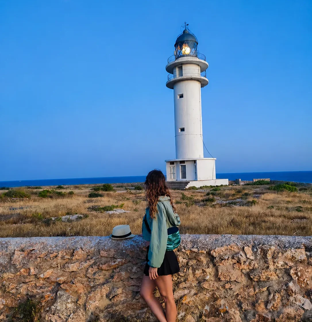 Mujer de pie junto a un faro blanco en un paisaje costero al atardecer, con una luna visible en el cielo azul. La mujer lleva una chaqueta ligera y una falda, y hay un sombrero sobre la pared de piedra.