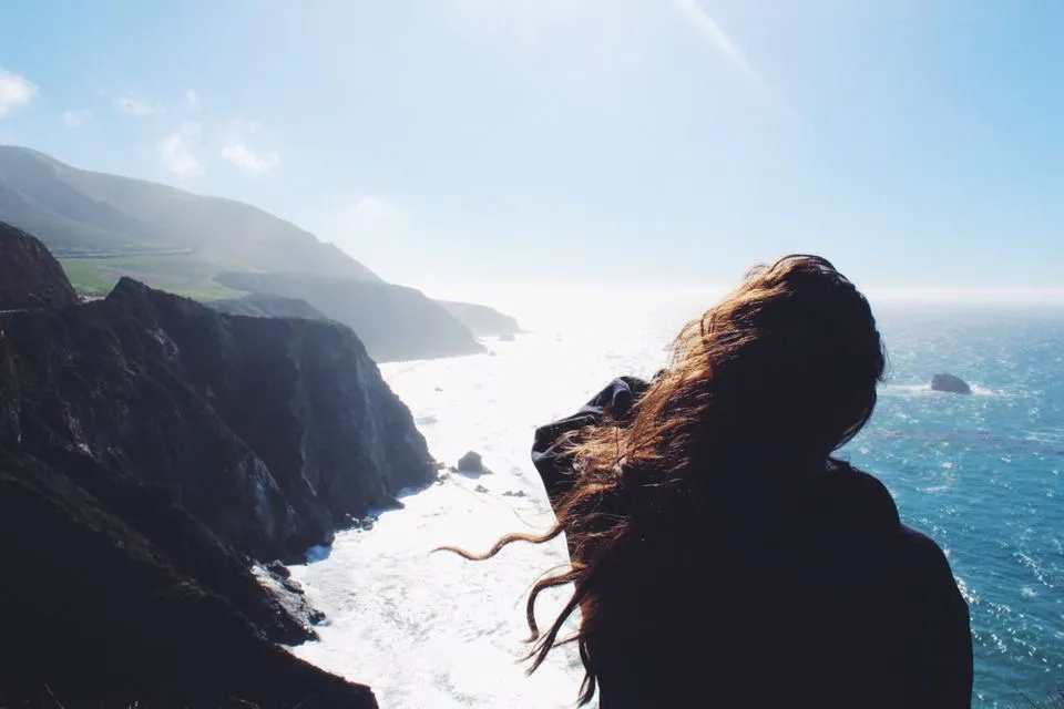 Mujer de espaldas disfrutando de un paisaje costero con acantilados y el mar en un día soleado. La brisa mueve su cabello mientras observa el océano.