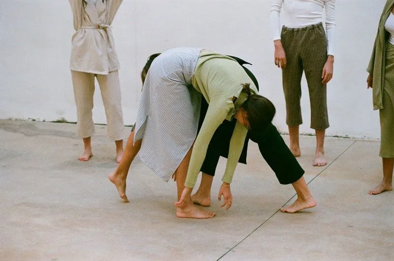 Grupo de bailarines en una coreografía contemporánea, mostrando una interacción dinámica. La escena se desarrolla en un espacio minimalista con un fondo blanco y un piso de cemento. Los bailarines están descalzos y visten ropa casual y elegante, enfatizando la expresión corporal y la conexión entre ellos.