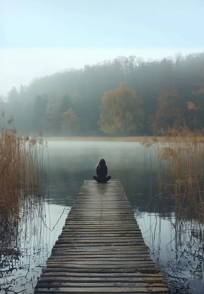 Mujer sentada en un muelle de madera sobre un lago tranquilo, rodeada de niebla y vegetación, creando una atmósfera serena y contemplativa.