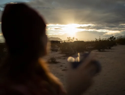 Mujer con gorro sosteniendo una taza mientras observa un atardecer en un paisaje desértico, con nubes y montañas al fondo.