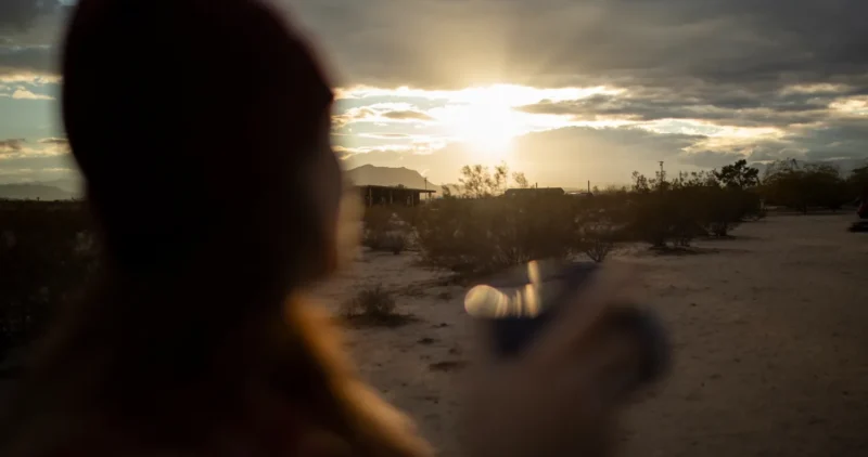 2149302406 Mujer con gorro sosteniendo una taza mientras observa un atardecer en un paisaje desértico, con nubes y montañas al fondo.