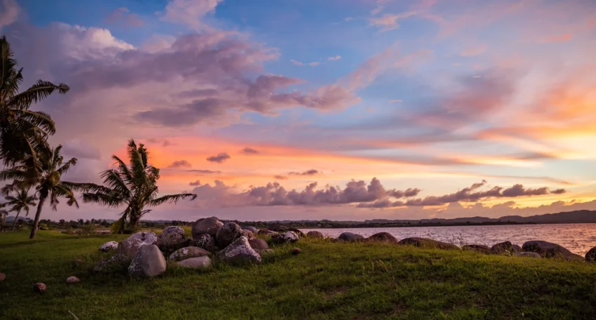 Atardecer sobre un paisaje costero con palmeras, rocas y un cielo lleno de nubes de colores cálidos en tonos naranjas y rosas.