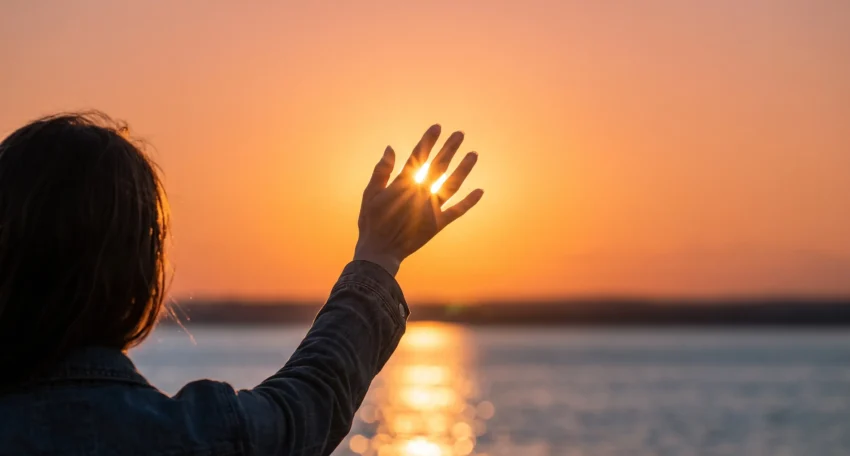 Persona levantando la mano hacia un atardecer sobre el agua, con luz del sol brillando entre los dedos, creando un ambiente sereno y reflexivo.
