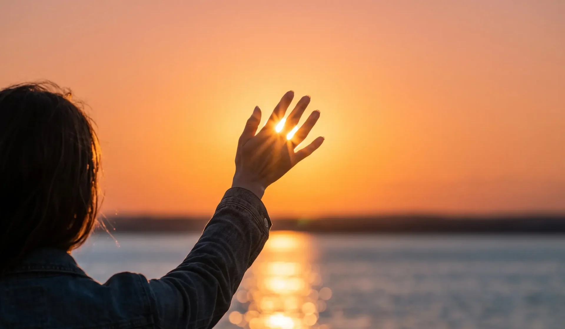 Persona levantando la mano hacia un atardecer sobre el agua, con luz del sol brillando entre los dedos, creando un ambiente sereno y reflexivo.