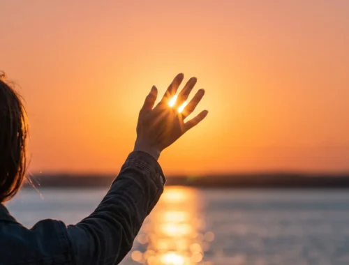 Persona levantando la mano hacia un atardecer sobre el agua, con luz del sol brillando entre los dedos, creando un ambiente sereno y reflexivo.